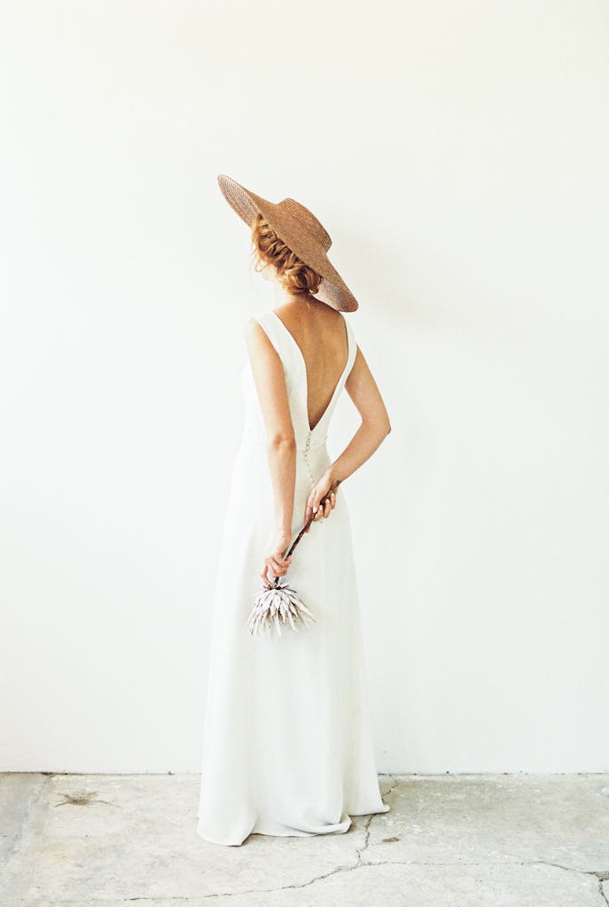 A stylish woman in a backless white dress and sun hat holds dried flowers, standing against a minimalist backdrop.