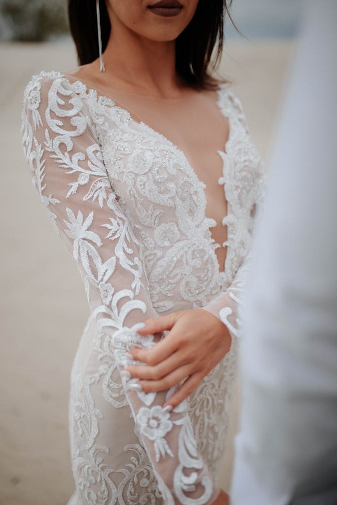 Bride in an elegant lace wedding dress standing on a sandy beach, showcasing intricate embroidery.